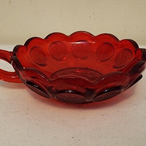 May include: A ruby red glass bowl with a handle. The bowl has a scalloped rim and a series of circular indentations around the edge. The handle is a simple loop shape. The bowl is likely vintage.