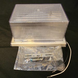 May include: A clear, rectangular outdoor light fixture with a ribbed, textured cover. The fixture sits on a white base, with a clear plastic bag containing mounting hardware. Electrical wires extend from the base.