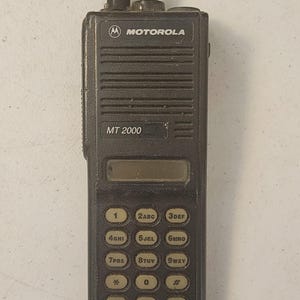 May include: A dark gray Motorola MT 2000 two-way radio. The radio has a keypad with numbers and letters, a small display screen, and an antenna. The Motorola logo is visible at the top of the device.