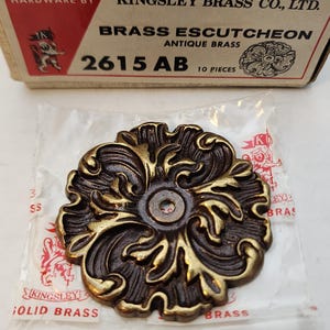 May include: A solid brass escutcheon with an antique brass finish, featuring an ornate floral design. The product is from Kingsley Brass Co., Ltd. and is labeled as cabinet hardware. The packaging includes the text "2615 AB" and "10 pieces."