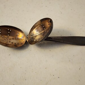 May include: A vintage silver tea infuser with an oval-shaped perforated bowl and a long handle. The infuser is open, revealing the interior. The handle has a decorative pattern. The tea infuser is designed to hold loose tea leaves.
