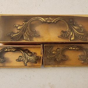 May include: Ornate, antique-style brass drawer pulls with a scrolling leaf design. The pulls are displayed in their original cardboard boxes with clear plastic covers. The hardware has a vintage aesthetic.