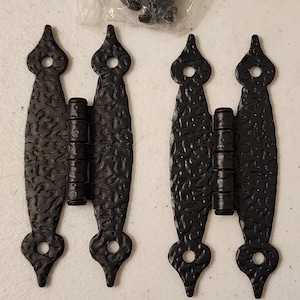 May include: Two black decorative hinges with a textured surface and a teardrop design. The hinges are made of metal and have a rustic appearance. A small bag of screws is visible in the background.