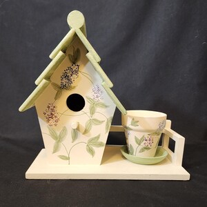 May include: A white birdhouse with a green roof and a small flower pot on a white base. The birdhouse and pot are decorated with a floral pattern.