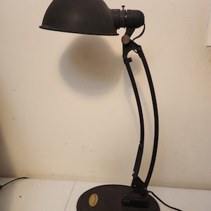 May include: A black desk lamp with a curved arm and a rounded shade. The lamp has a circular base and a flexible arm for adjusting the light's direction. The lamp is made of metal and has a vintage appearance.