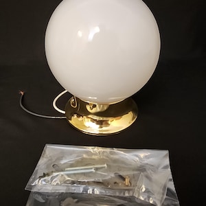 May include: A white globe light fixture with a gold-colored base. The fixture has a round, opaque glass shade and a brass-colored base. A package of hardware is included. The light fixture is on a black surface.