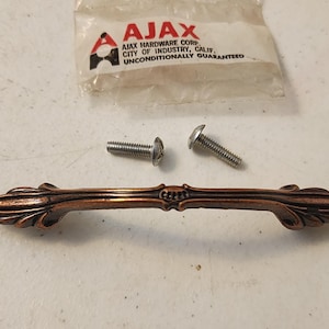May include: A bronze-colored cabinet pull with ornate floral detailing, accompanied by two silver screws and a small plastic package with the "AJAX" logo and text. The pull is approximately 6 inches long.