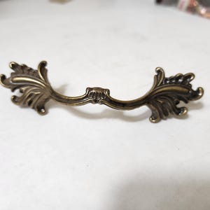 May include: A decorative brass drawer pull with a floral design. The pull is shaped like a curved arch with ornate details on each end.