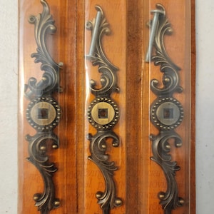 May include: Three antique bronze-colored decorative hardware pieces. Each piece has an ornate, scrolling design with a central circular medallion and a square center. The hardware is packaged in clear plastic with a brown backing.