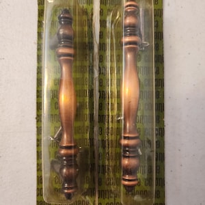 May include: Two bronze-colored cabinet handles in clear plastic packaging. The handles have a decorative, turned design with a dark brown finish on the ends. The packaging has the word "colonnade" printed on it.