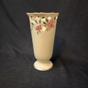 May include: A white ceramic vase with a delicate floral design and a scalloped rim. The vase has a gold trim around the top and bottom.