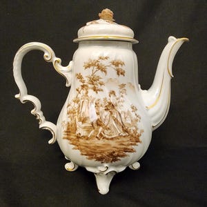 May include: White porcelain teapot with a brown pastoral scene, featuring a lid with a small knob and a curved handle. The teapot has a spout with gold trim and sits on three small feet. A decorative piece for serving tea.