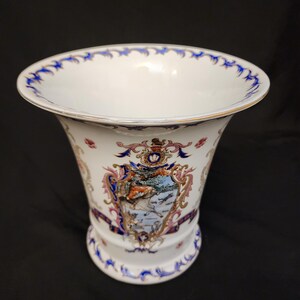 May include: White porcelain vase with a flared rim and a detailed crest design. The vase features blue and gold trim, with a decorative crest in the center. The crest includes a landscape scene with a horse and rider.
