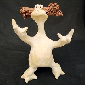May include: A whimsical, off-white ceramic figurine with outstretched arms and legs. The figure has a long neck, large eyes, and brown, textured hair. The sculpture has a slightly aged appearance, suggesting a vintage or handmade quality.