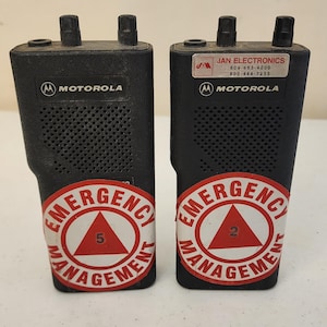 May include: Two black Motorola two-way radios. Each radio has a speaker grill, two antennas, and a red and white Emergency Management sticker. One sticker has a "5" and the other has a "2".