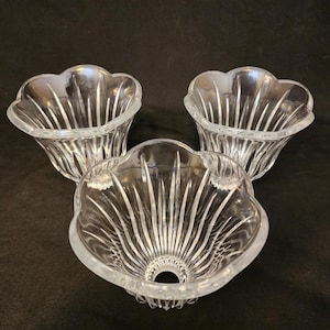 May include: Three clear glass decorative bowls with a scalloped edge and vertical etched lines. The bowls are arranged on a dark background. The bowls have a flower-like shape.