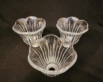 May include: Three clear glass decorative bowls with a scalloped edge and vertical etched lines. The bowls are arranged on a dark background. The bowls have a flower-like shape.