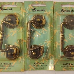 May include: Three antique brass cabinet pulls in their original packaging. Each pull has a decorative handle and is packaged on a green card with the Amerock logo and the text "A-876-AE ANTIQUE ENGLISH".