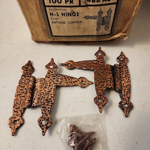 May include: Antique copper H-L hinges with a textured finish, alongside a packet of matching screws. The hinges are part of the Colonial Trim series, as indicated on the packaging. The packaging also indicates the finish is antique copper.