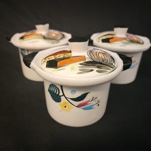 May include: Three white ceramic lidded pots with black handles. The lids are decorated with food illustrations, including cheese and onions. The pots have floral designs on the sides. The background is black.