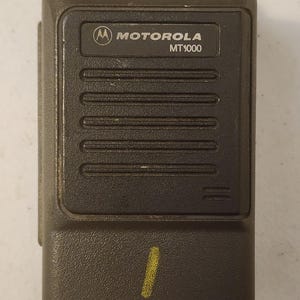 May include: A dark gray Motorola MT1000 two-way radio. The radio has two black knobs on top and a speaker grill. The Motorola logo and model name are printed on the front. A yellow mark is visible on the lower part of the radio.