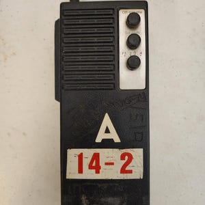 May include: A black Motorola two-way radio with a silver control panel and three black knobs. The radio has a label with the text "A" above "14-2" in red. The Motorola logo and "HANDIE-COM" are visible at the bottom.
