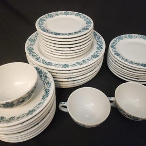 May include: A collection of white ceramic dinnerware with a teal floral pattern. The set includes plates of various sizes and teacups. The dishes are stacked in neat piles, showcasing a classic and elegant design.
