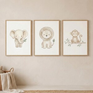 Op de afbeelding: Drie ingelijste aquarelprints met een baby-olifant, leeuw en aap. De olifant is lichtgrijs en houdt een groen blad vast. De leeuw heeft een bruine manen en staart. De aap zit op een tak. Elke print zit in een lichtbruine lijst.