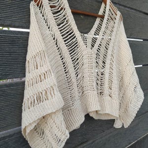 Pode incluir: Top de crochê creme de tecido aberto, com decote em V profundo e mangas curtas. O top está pendurado em um cabide de madeira contra um fundo de madeira cinza escuro. O design de crochê apresenta trabalhos abertos verticais e horizontais.
