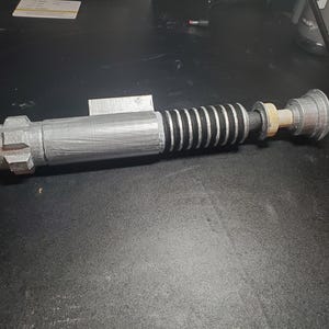 May include: A silver and black lightsaber hilt prop featuring a textured metallic cylindrical body, a ribbed black grip section, a small rectangular control box on the side, and a flared pommel at the end with a light-coloured circular accent piece.