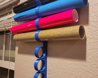 May include: A wall-mounted vinyl roll holder in blue, holding rolls of black, blue, pink, and gold glitter vinyl. The holder is mounted on a beige wall near a window with blinds. The holder has a series of blue rings to hold the rolls.