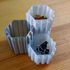 Puede incluir: Tres contenedores hexagonales de almacenamiento en plata y blanco. Los contenedores están apilados. Uno contiene chinchetas de colores, otro pinzas para papel negras y el tercero está vacío. Los contenedores son de material acanalado.