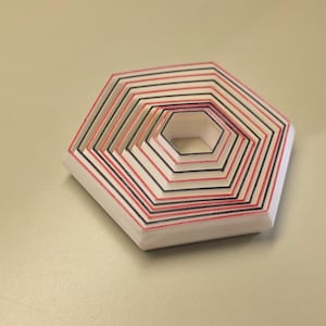 May include: A white, multi-layered, hexagonal paper craft with red and black stripes. The nested design creates a three-dimensional effect. The object is resting on a light beige surface.