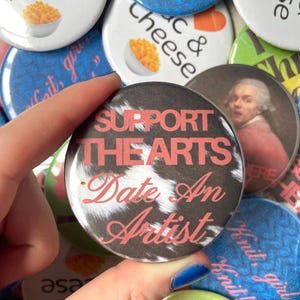 May include: A round pinback button featuring a black and white cow print background with the text "SUPPORT THE ARTS" in bold red capital letters and "Date An Artist" in a red cursive script font, centred on the surface of the circular pin.