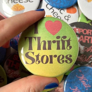 Peut inclure: Un badge rond avec un fond vert, un cœur rouge et le texte brun foncé "I Thrift Stores" imprimé au centre. Ce badge est entouré d'une collection d'autres épinglettes circulaires présentant divers motifs et inscriptions textuelles.
