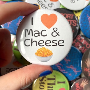 May include: A round white pin-back button featuring the black text I Mac & Cheese, an orange heart icon, and a small illustration of a bowl of macaroni and cheese. The button is surrounded by a collection of various other colourful pin-back buttons.