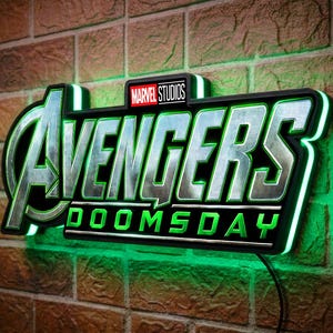 May include: A wall-mounted LED sign featuring the metallic silver Avengers logo and the green text DOOMSDAY, topped with a small Marvel Studios logo. The sign emits a bright green glow from behind its black frame, creating a vibrant light effect.