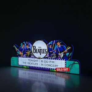 Op de afbeelding: Een blauw en wit bord met de tekst "The Beatles" in een cirkel. Het bord heeft ook een afbeelding van de Beatles die muziek maken. Het bord zegt "Tonight - 8:00 P.M. The Beatles - In Concert" en "Sold Out".