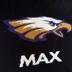 Könnte beinhalten: Schwarzer Strickstoff mit einem Häkeldesign eines Adlerkopfes in Weiß, Gold und Lila. Das Wort "MAX" wird in weißen Blockbuchstaben unter dem Adler angezeigt.