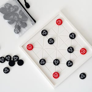 Puede incluir: Un juego de mesa blanco con un diseño geométrico, con piezas de juego negras y rojas. Las piezas negras tienen un gráfico blanco y las piezas rojas tienen un gráfico blanco. También se incluye una bolsa de malla con piezas adicionales.