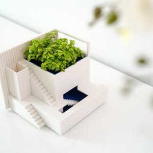 Puede incluir: Un macetero de cerámica blanca con forma de casa moderna con una planta verde creciendo en el techo. El macetero tiene una pequeña piscina azul y escaleras que conducen al techo.