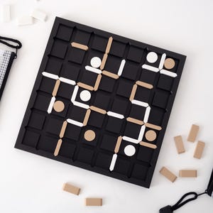 May include: A black wooden board game with a grid pattern, featuring white and tan game pieces. The game is set on a white surface, with small tan and white pieces scattered around. Two mesh zipper pouches are also present.