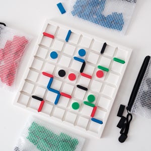May include: A white square board game with a grid pattern, featuring colorful game pieces in red, blue, green, and black. The game is accompanied by zippered mesh bags containing additional pieces. A fun, colorful board game.