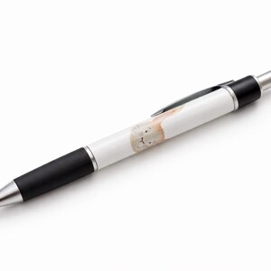 Op de afbeelding: Een witte balpen met een zwarte grip en zilveren accenten. De pen heeft een lichtbruine illustratie van een konijn op de schacht. De pen staat schuin op een witte achtergrond.