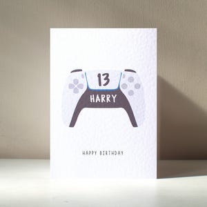 Puede incluir: Tarjeta de cumpleaños blanca con una ilustración de un mando de videojuegos. El mando tiene botones grises, el número 13 y el nombre HARRY. Las palabras FELIZ CUMPLEAÑOS están impresas debajo. Una tarjeta divertida para un adolescente.