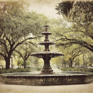 Op de afbeelding: Een foto in vintage-stijl van een gelaagde fontein in een park. De fontein is gemaakt van donker metaal en heeft water dat uit de niveaus stroomt. Weelderige bomen met Spaanse mos omlijsten de fontein en creëren een serene en pittoreske scène.