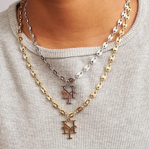 May include: Two layered necklaces with a gold and silver finish. Each necklace features a chain with oval links and a pendant with the letters "NY" in a stylized design. The necklaces are displayed against a gray background.