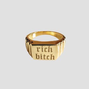 May include: A gold-colored ring with the words "rich bitch" engraved in a gothic font. The ring has a rectangular face and a ribbed band, set against a neutral background.