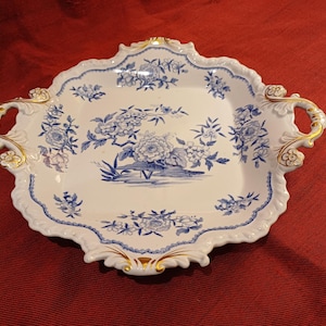 以下が含まれることがあります： 青い花柄と金色のアクセントが施された白い磁器製のサービングトレイ。トレイはスカラップエッジと装飾的なハンドルを備えています。花柄には牡丹が含まれています。サービング用の装飾品。