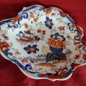 May include: A white, scalloped-edge decorative plate with a floral and pagoda design. The plate features orange, blue, and red floral patterns and a blue rim. The plate is likely made of porcelain or ceramic.